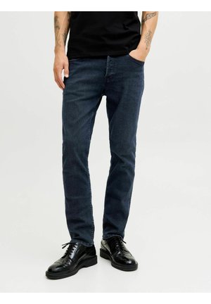 Jeans slim-fit de culoare albastru închis, cu o textură subtilă, buzunare frontale, purtate cu pantofi din piele negri cu detalii tip brogue. Se vede un tricou negru.