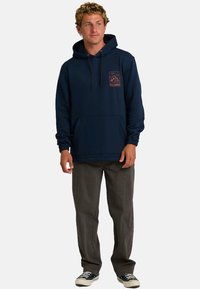 Młody mężczyzna w granatowym hoodie z grafiką na klatce piersiowej, w szarych spodniach i czarnych trampkach, stojący na jednolitym białym tle.