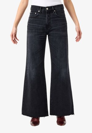 Persona che indossa jeans di jeans neri a vita alta con gamba larga, orli grezzi e scarpe a punta di colore rosso scuro su sfondo bianco.