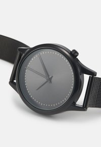 Kulaté černé hodinky s minimalistickým designem, šedým ciferníkem, stříbrnými číslicemi a černým síťovým páskem. Na boku mají černou korunku.