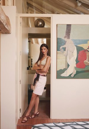 Femme portant un haut fleuri et une jupe rose, debout dans une porte avec les bras croisés, à côté d'une grande peinture abstraite sur un mur blanc.