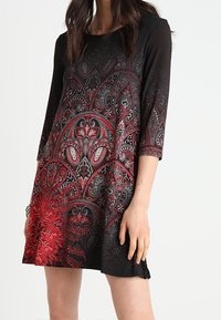 Czarna sukienka z czerwono-srebrnym ozdobnym nadrukiem w motyw paisley. Posiada rękawy do połowy przedramienia i luźny krój, z lekko rozszerzanym dołem.