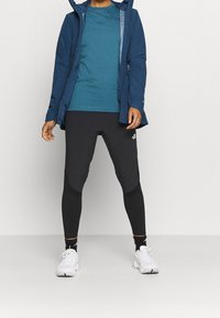 Niebieska kurtka z kapturem, turkusowa koszulka, czarne legginsy z logo, białe sneakersy. Nowoczesny design sportowy, dopasowany, odpowiedni do aktywności na świeżym powietrzu.