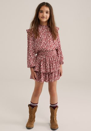 Röd blommig klänning med lång ärm, volangdetaljer och en lager-på-lager kjol. Tillsammans med bruna western-stil stövlar och randiga strumpor.