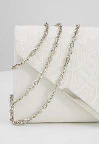 Pochette blanche avec superposition en dentelle, bandoulière chaîne argentée, rabat triangulaire avec accent métallique. Texture lisse avec motif de dentelle complexe.