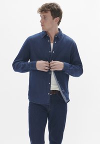 Camisa de denim, azul oscuro, diseño con botones, corte regular, mangas largas, textura suave, combinada con una camiseta clara debajo y jeans oscuros.