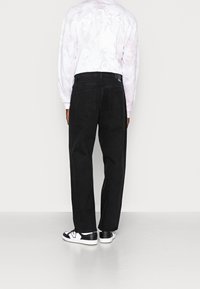 Osoba w czarnych prostych dżinsach, białych i czarnych trampkach oraz jasnej koszuli z długim rękawem z delikatnym nadrukiem z tyłu, stoi.