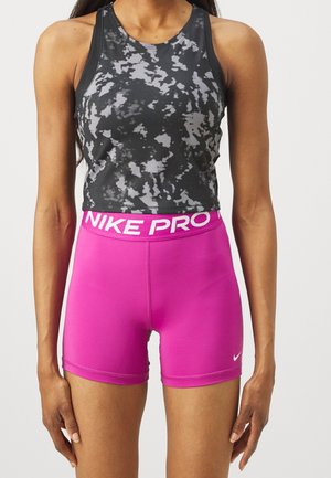 Frau trägt ein ärmelloses schwarz-graues Batik-Cropped-Top und leuchtend pinke Nike Pro Sportshorts, steht mit entspannten Armen.