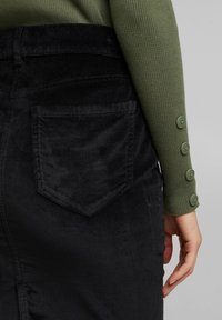 Gros plan d'une personne portant un pantalon en velours côtelé noir et un haut vert côtelé à manches longues avec quatre boutons sur le poignet, main détendue sur le côté.