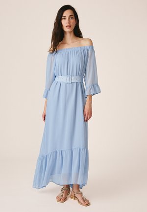 Woman wearing a light blue off-shoulder maxi dress with a matching belt and ruffled hem, paired with embellished flat sandals.