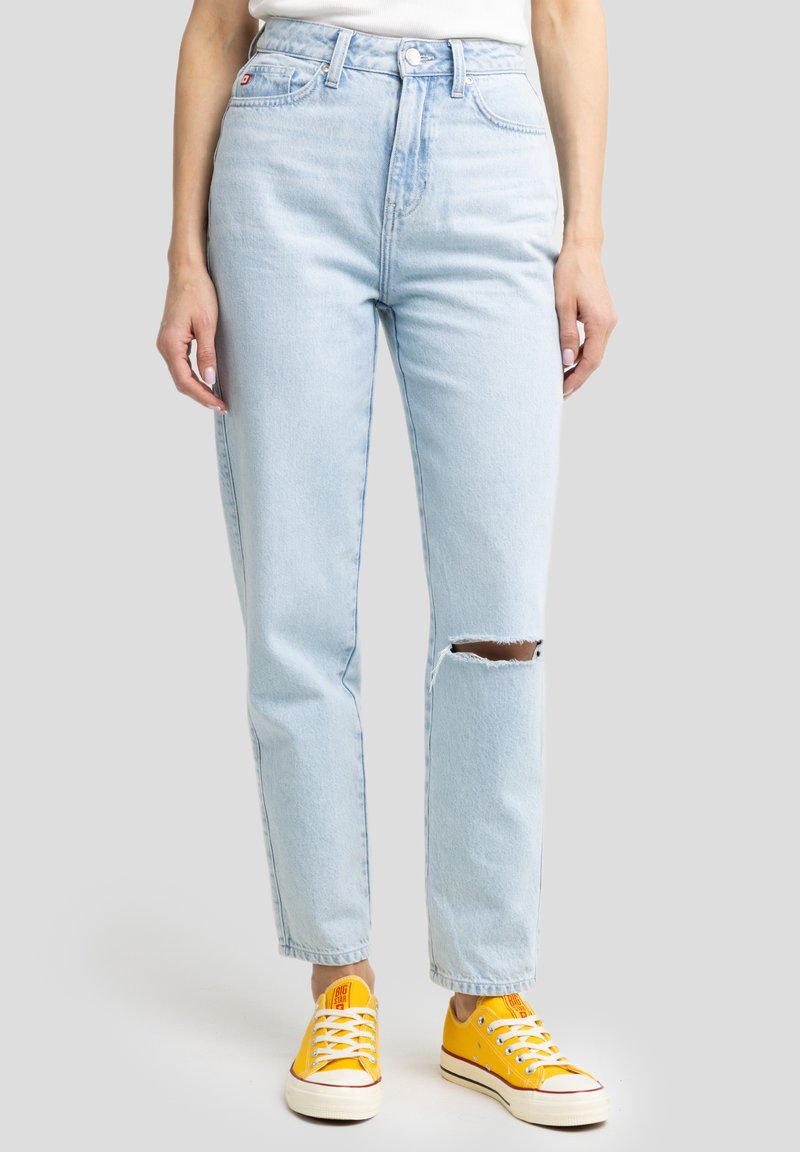 Vaqueros de tiro alto de color azul claro con un corte en la rodilla, hechos de mezclilla, combinados con zapatillas de lona amarillas. Diseño de pierna recta y frente plano.