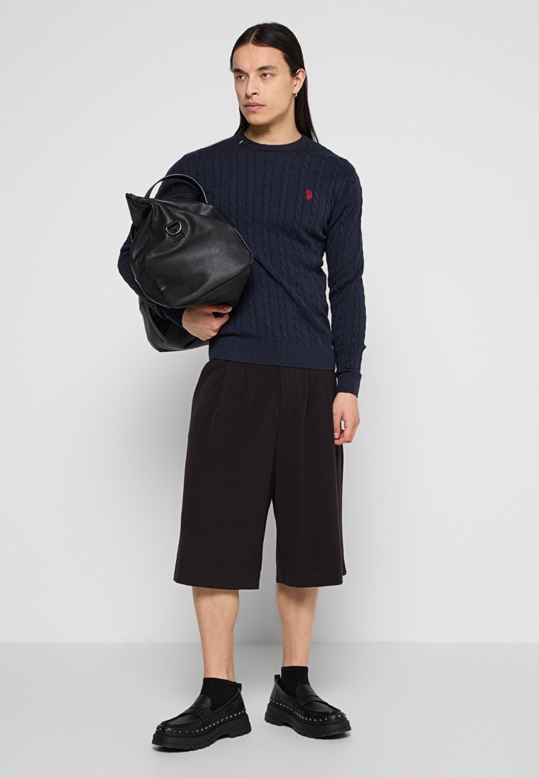 Marineblauer Kabelstrickpullover, schwarze gefaltete Shorts, schwarze oversized Lederhandtasche und klobige schwarze Schuhe mit Nietenakzenten.