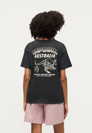 Άτομο με σγουρά μαλλιά φοράει μαύρο t-shirt Rip Curl Australia με γραφικό σχεδιασμό καγκουρό και σανίδας του σερφ, συνδυασμένο με ροζ υφασμένα σορτς.