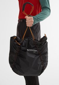 Schwarze Nylontasche mit geraffter Textur, ausgestattet mit orangefarbenen und braunen Kordeln sowie einem Emblem-Label. Abgerundetes Design mit weichen Griffen.