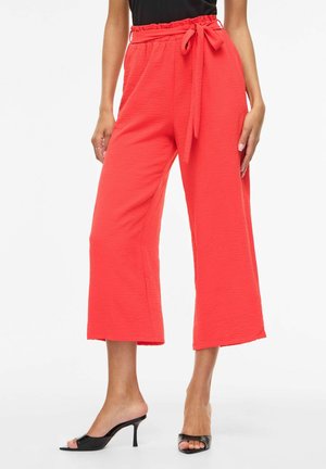Kvinde iført lyse koralfarvede, vidde, cropped bukser med bindebælte i taljen og sorte åbne hæle.