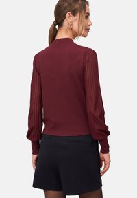 Femme aux cheveux bruns en queue de cheval, portant un haut bordeaux avec des manches bouffantes transparentes et un short noir, vue de dos.