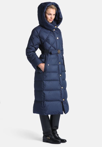 Femme portant un long manteau matelassé bleu marine avec capuche, taille cintrée avec ceinture, boutons dorés, mains dans les poches, associé à des bottes noires.