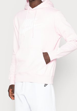 Osoba ubrana w jasnoróżową bluzę z kapturem Nike z białym logo i białe spodnie dresowe Nike, stoi z jedną ręką w kieszeni bluzy.