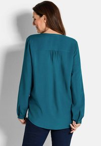 Blusa lunga a maniche con girocollo in teal, con dettaglio a plissè sul retro e polsini con bottoni. Tessuto morbido con una texture liscia e schiena allungata.