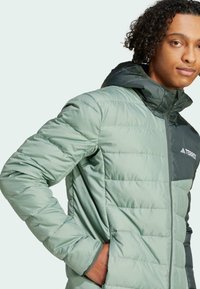 Chaqueta acolchada en verde claro con capucha verde oscura. Presenta un cierre frontal de cremallera, puños elásticos y bolsillos laterales. Incluye un logotipo en el pecho.