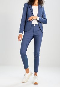 Modrý oblekový blejzr s ladícími fitted kalhotami, vyrobený z hladkého materiálu. Kombinovaný s bílými botami, které mají minimalistický design a nenápadné knoflíky.