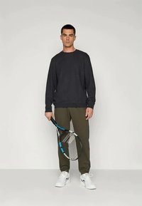 Maglietta grigia scura a maniche lunghe abbinata a pantaloni verde oliva, con una racchetta da tennis in mano e indossando sneaker bianche. Stile semplice e atletico.