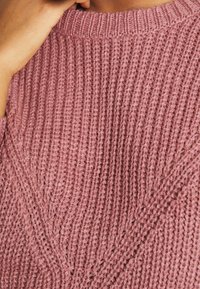 Gebreide trui in gedempt roze, met ribbelstructuur. Close-up toont een diagonaal patroon en zachte, dikke vezels. Ronde halslijn.