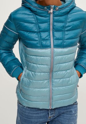 Person wearing blue two-tone padded jacket with hood and front zipper, hands in pockets, paired with blue jeans.