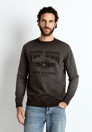 Mand med mørkt krøllet hår og skæg iført en sort Top Gun sweatshirt og blå jeans, stående mod en ensfarvet lys baggrund.