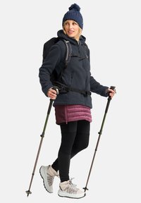 Fleece jack in donkerblauw, geïsoleerde bordeauxrode rok, zwarte leggings en lichtgrijze wandelschoenen. Draagt trekkingstokken en een rugzak.