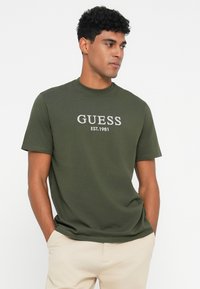 Olijfgroene katoenen t-shirt met korte mouwen, ronde hals en geborduurd "GUESS EST.1981" in het wit aan de voorkant. Gladde textuur.