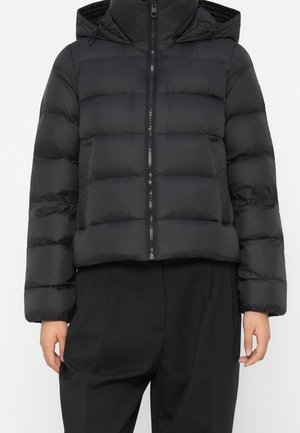 Veste matelassée noire avec fermeture éclair à l'avant et capuche, portée avec un pantalon noir, mains relâchées le long du corps.