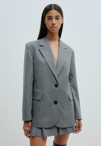 Grau blazer mit taillierter Passform, zwei schwarzen Knöpfen, Reverskragen und seitlichen Taschen, ergänzt durch einen plissierten Rock darunter.