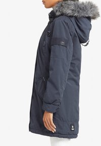 Námořnicky modrá parka s kapucí lemovanou umělou kožešinou, zipovými kapsami, elastickými manžetami a značkovou nášivkou na lemu. Hladká textura látky.