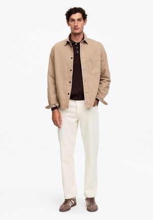 Mężczyzna stoi, ubrany w beżową kurtkę zapinaną na guziki na jasnobrązową koszulkę polo, białe spodnie i brązowe trampki z białymi paskami, jednolite białe tło.