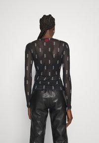 Langärmliges schwarzes Mesh-Top mit wiederholtem weißen Logo-Muster; kombiniert mit hochgeschnittenen schwarzen Lederhosen. Rotes Etikett am Hals.