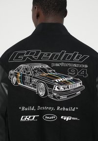 Fekete dzseki, amelyen egy hímzett versenyautó grafika látható színes csíkokkal. Tartalmazza a következő szövegeket: "GRedy teljesítmény 94" és "Építsd, pusztítsd el, építsd újjá."