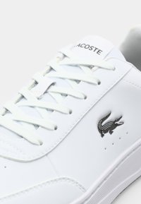 Baltos odinės sportinės bateliai su apvaliu pirštu, plokščiomis raišteliais ir juodu Lacoste krokodilo logotipu šone. Lygi tekstūra ir minimalistinis dizainas.