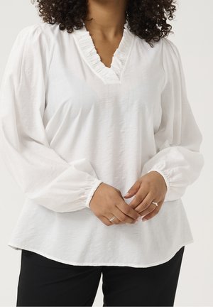 Kvinde med krøllet hår iført en hvid bluse med lange ærmer og flæsekrave, sorte bukser og ringe på fingrene.