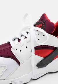 Witte sneaker met bordeauxrode en zwarte accenten. Beschikt over een gestructureerde leren bovenwerk, gewatteerde kraag en witte veters. Logo op de tong.