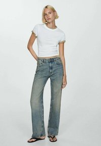 T-shirt bianco corto con bordi beige abbinato a jeans azzurri svasati e sbiaditi. Modella indossa sandali neri, in piedi contro uno sfondo neutro.