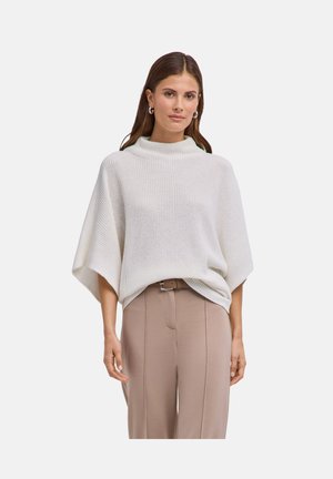 Femme portant un pull poncho en maille blanche et un pantalon beige taille haute avec ceinture, debout devant un fond clair uni.