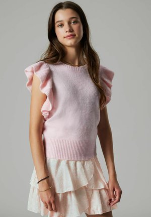 Ung kvinde med langt brunt hår iført en lyserød flæset ærmeløs sweater og et lagdelt blege lyserødt skørt mod en grå baggrund.