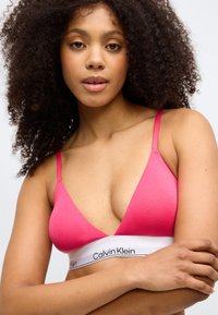 Rozā kokvilnas bralette ar dziļu V-veida kakla izgriezumu, plānām regulējamām siksniņām un baltu joslu ar melnu logo. Gluda tekstūra, minimālistisks dizains.