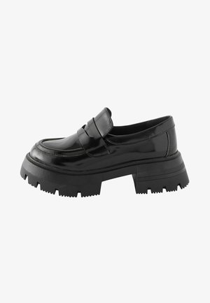 Čierne lakované kožené mokasíny s robustnou, textúrovanou čiernou gumovou podrážkou a dekoračným pásikom cez prednú časť. Hladký a lesklý povrch.