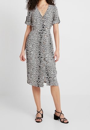 Kobieta ubrana w czarno-białą sukienkę midi w cętki lamparta z krótkimi rękawami oraz czarne sandały na obcasie, stojąca na jednolitym tle.