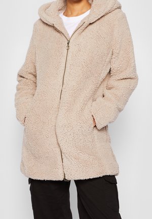 Personne portant une veste beige à texture douce avec capuche et fermeture éclair, les mains dans les poches, associée à un pantalon noir et une chemise blanche en dessous.