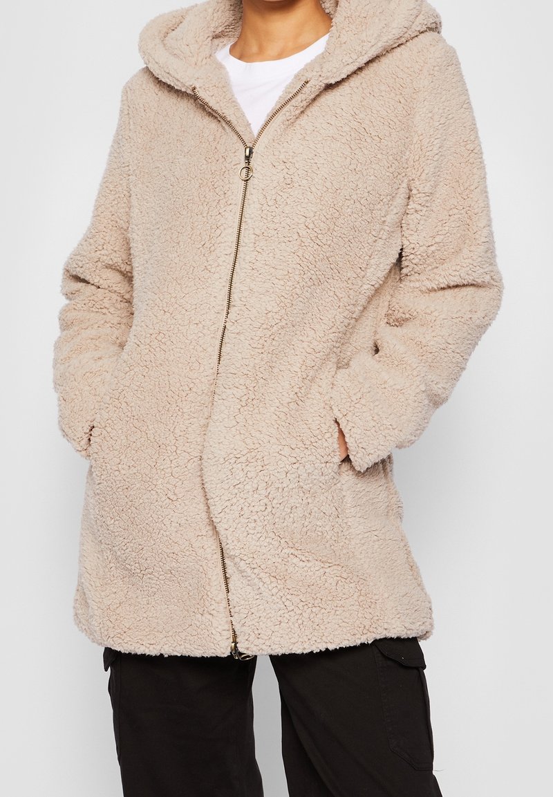 Personne portant une veste beige à texture douce avec capuche et fermeture éclair, les mains dans les poches, associée à un pantalon noir et une chemise blanche en dessous.