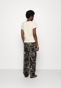 Camiseta de color beige claro con mangas cortas, combinada con pantalones de pierna ancha negros con contornos florales blancos. Textura suave y ajuste relajado.