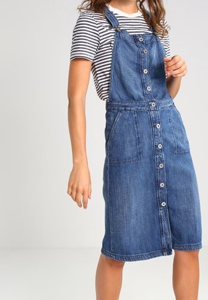 Kvinde iført blå denim pinafore-kjole med knapper foran over stribet kortærmet skjorte, stående mod en ensfarvet lys baggrund.